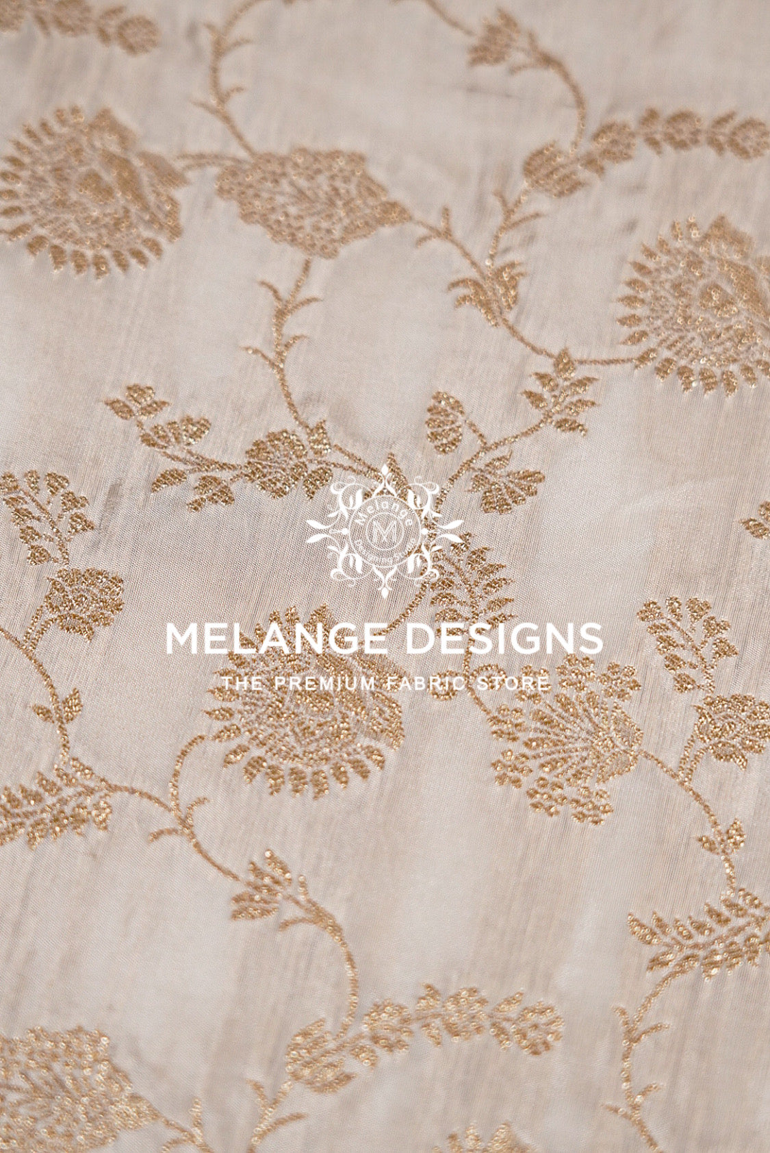 A close-up of a light beige Silk Brocade Dyeable Fabric with elegant metallic gold floral embroidery.