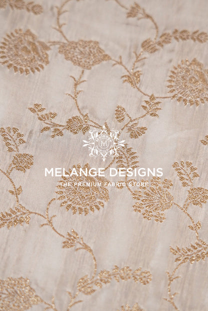 A close-up of a light beige Silk Brocade Dyeable Fabric with elegant metallic gold floral embroidery.