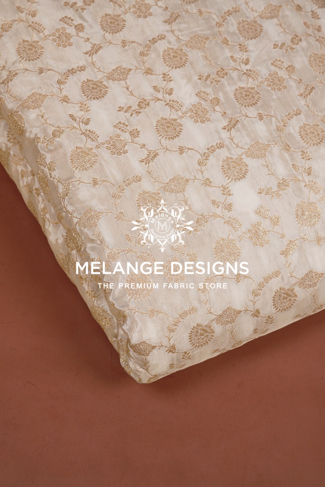 A close-up of a cream-colored Silk Brocade Dyeable Fabric with a beige floral jacquard pattern, folded diagonally over a solid terracotta brown background. 