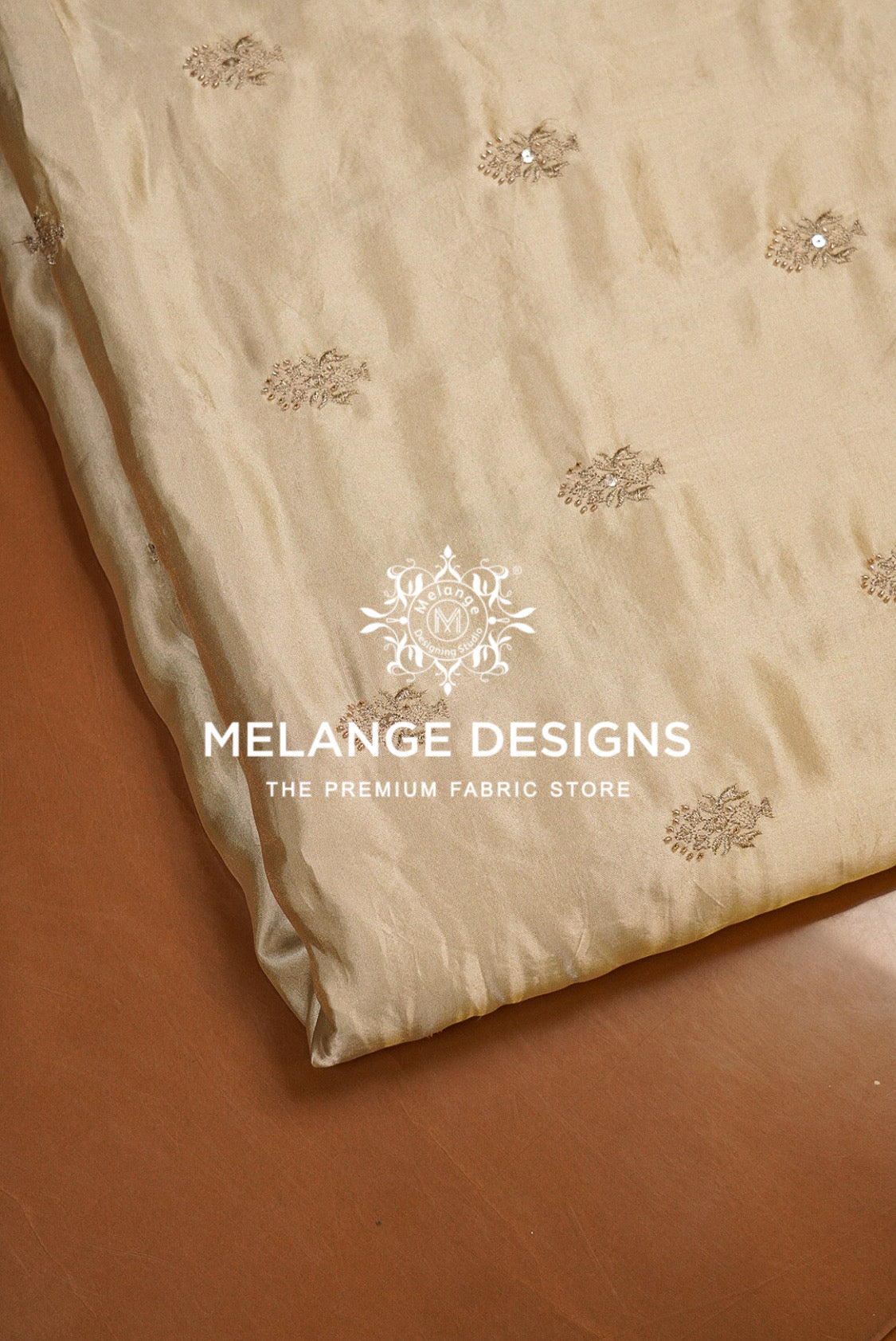 A folded beige Tissue Satin Embroidery Fabric Dyeable with delicate gold floral embroidery and small sequins, displayed on a brown surface. 