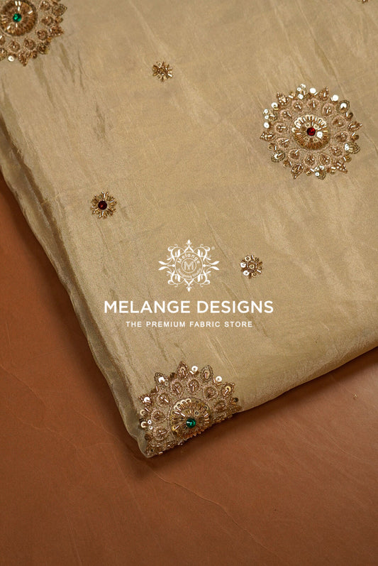 A close-up of a beige Tissue Silk Embroidery Fabric with ornate gold and silver embroidery, including floral motifs and small red and green accents.